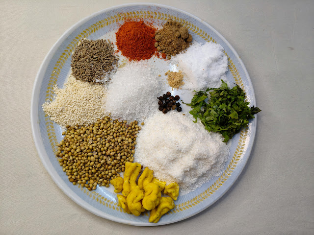 Assorted Indian spices including cumin seeds, coriander seeds, red chili powder, salt, coconut, turmeric, and fresh herbs arranged on a plate